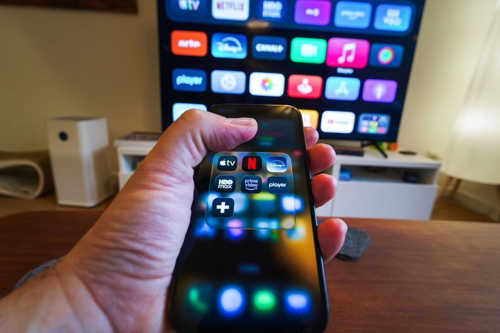 Mão segurando um celular com ícones de aplicativos de streaming em frente a uma televisão