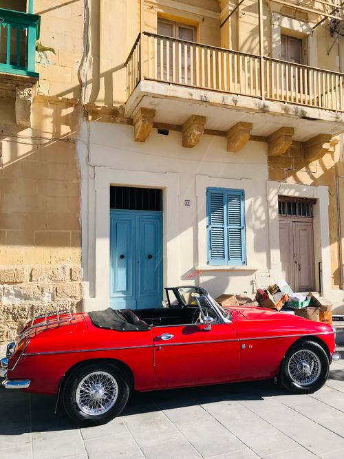 Free A classic red convertible car parked against a historic building façade on a sunny day. Stock Photo