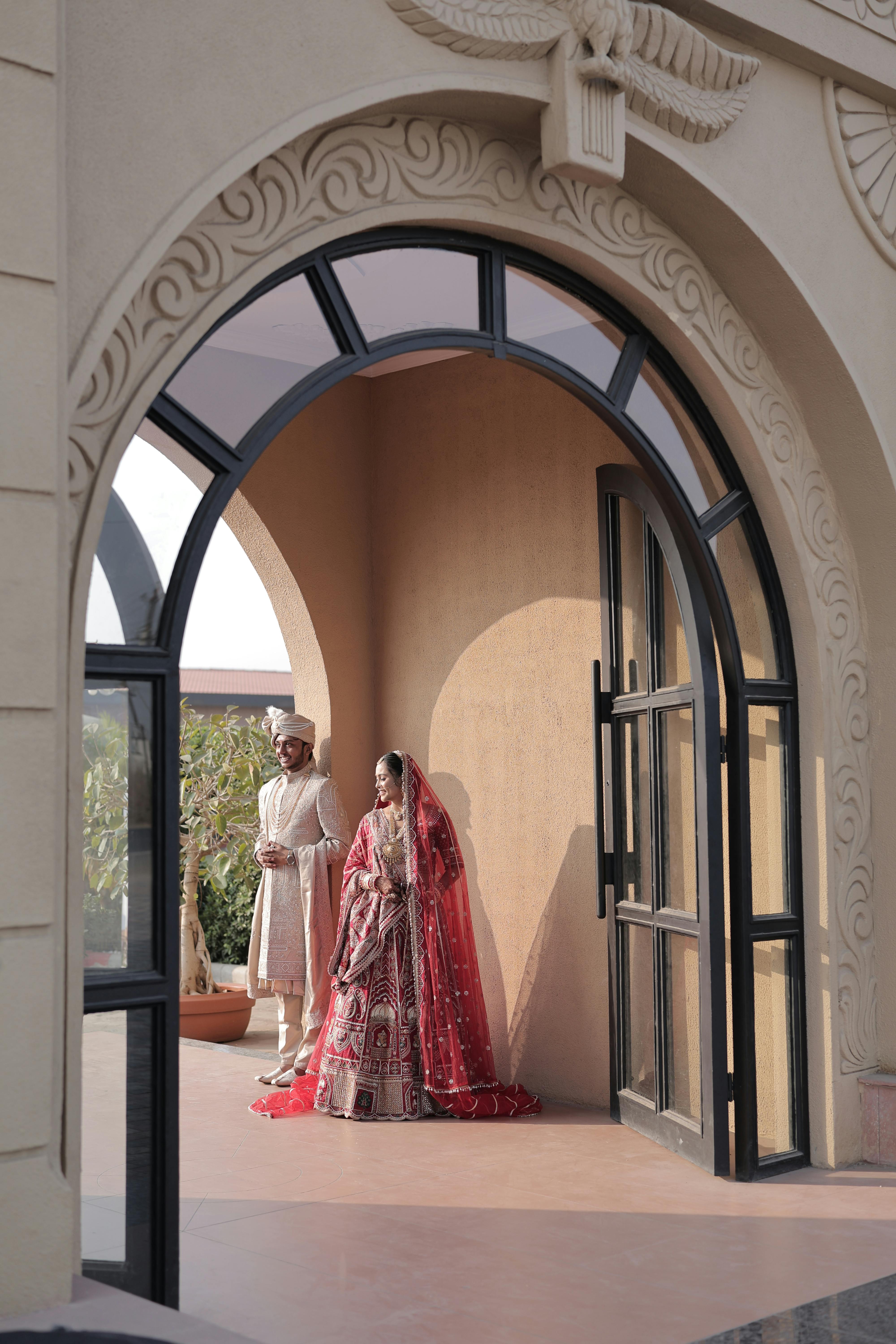 Gratis Novia y novio con atuendo tradicional bajo un arco ornamentado, creando una escena de boda elegante y atemporal. Foto de stock