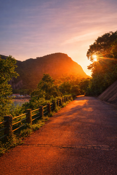 A serene path illuminated by vibrant sunset hues with mountains and lush greenery.