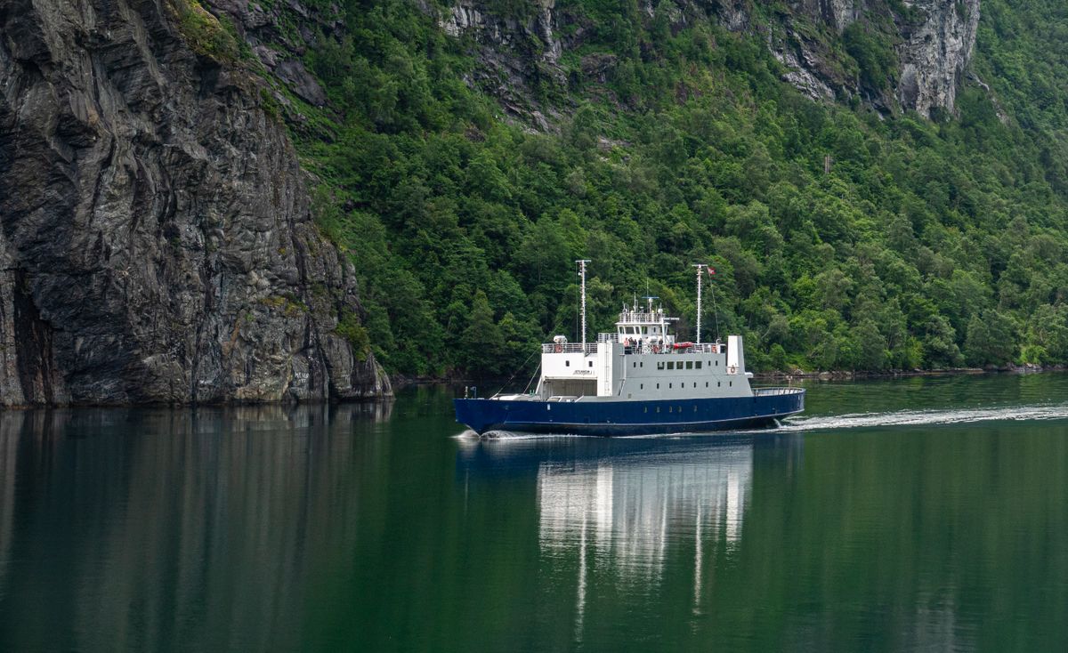 A small blue-and-white ferry crossing a narrow fjord between steep, forested green cliffs, the water calm and mirroring the hillsides.