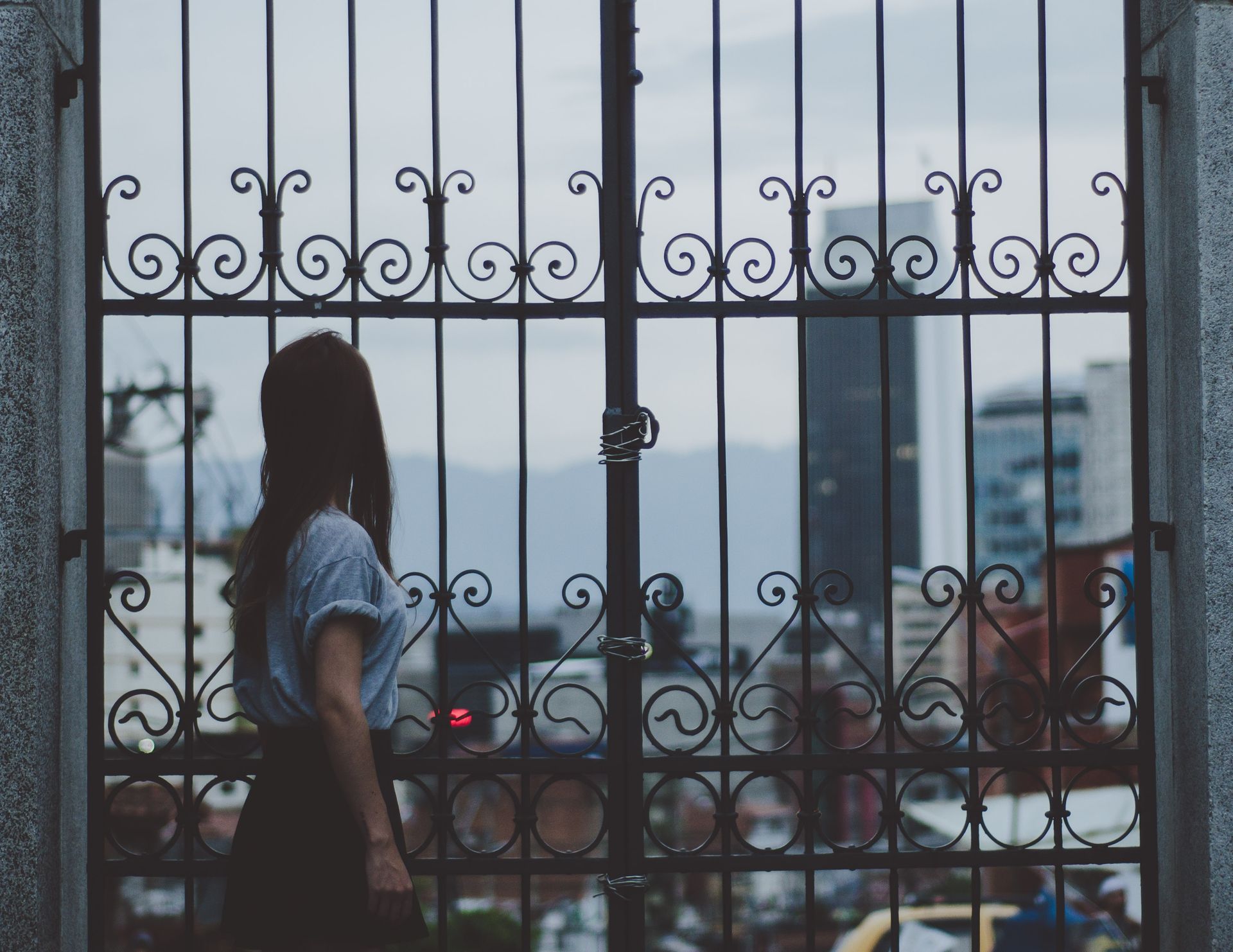 A woman gazes through a decorative iron gate at an urban cityscape, evoking a sense of nostalgia.