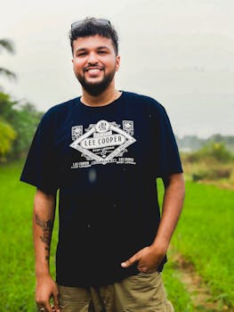 Man standing in a lush green field, smiling confidently with palm trees in the background.