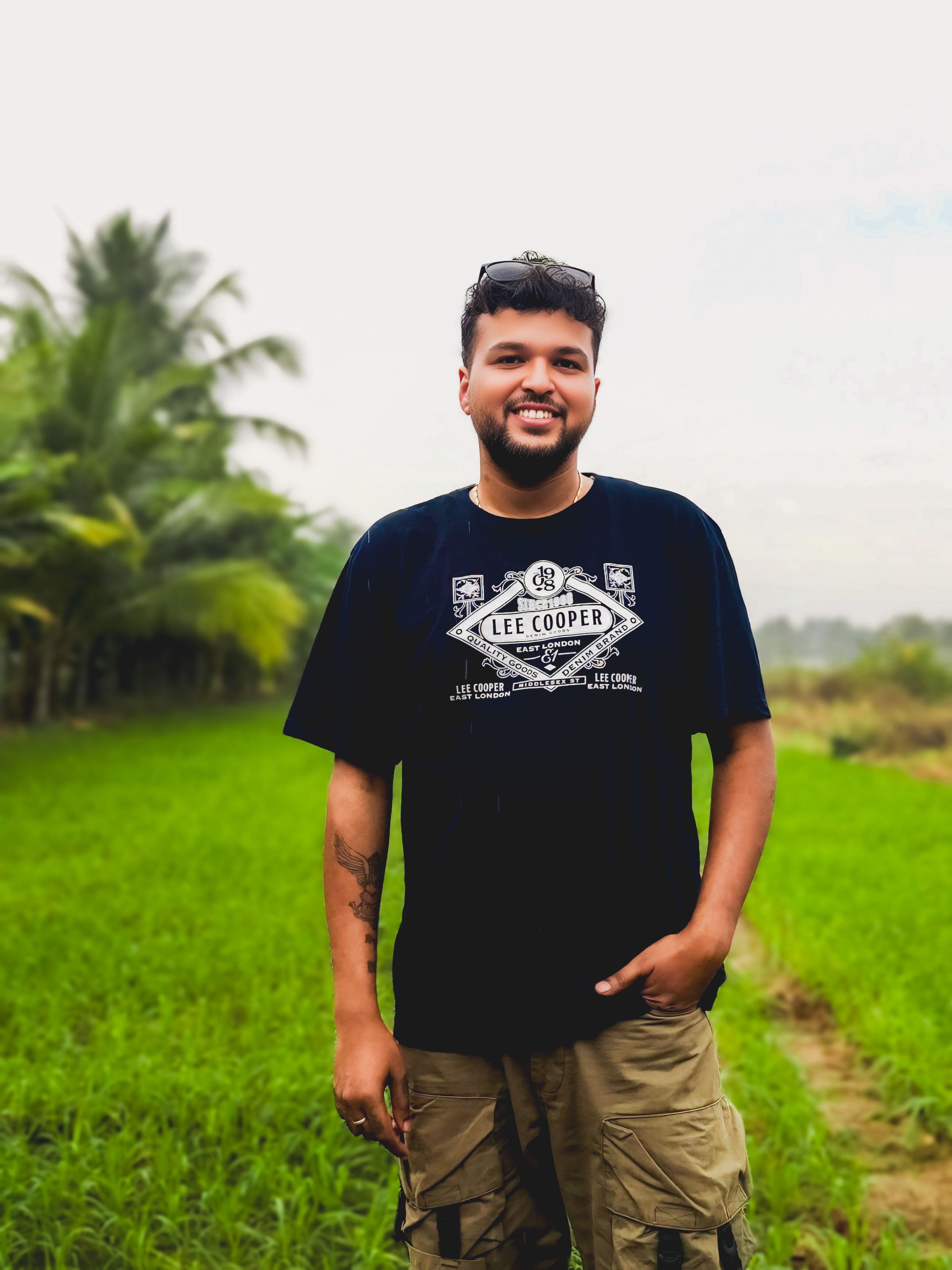A cheerful man stands in a vibrant green field, wearing a black T-shirt and cargo shorts.