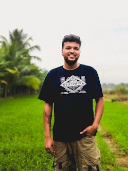 A cheerful man stands in a vibrant green field, wearing a black T-shirt and cargo shorts.