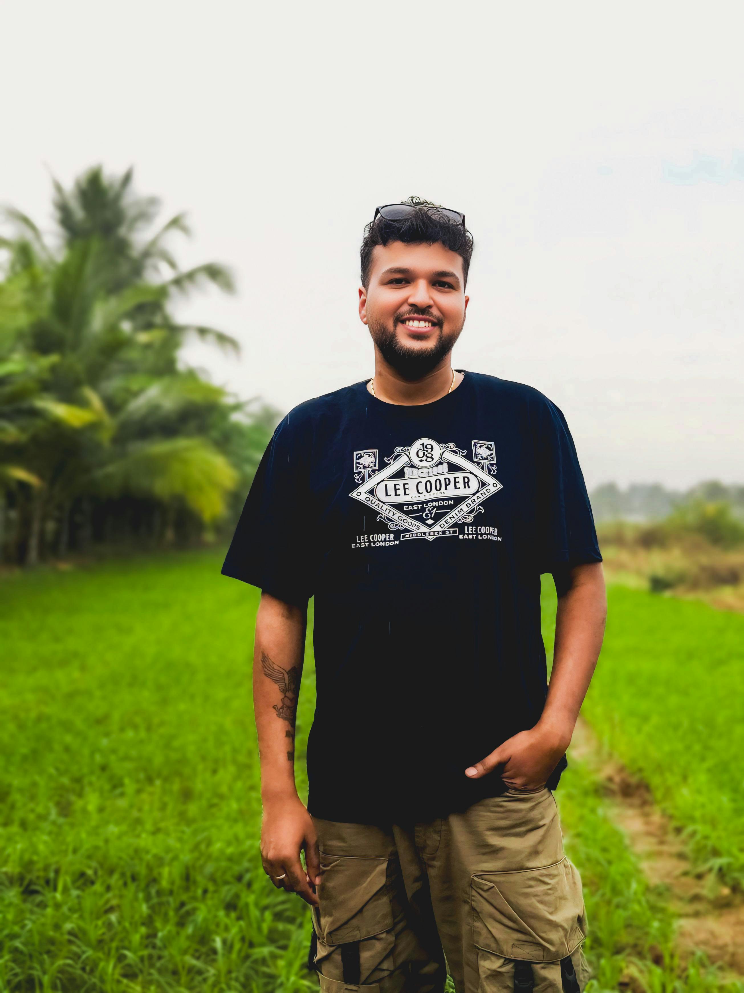Gratis Un hombre alegre se encuentra en un campo verde vibrante, vistiendo una camiseta negra y pantalones cortos cargo. Foto de stock