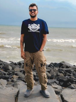 A man in casual attire stands on rocky seashore, with waves crashing in the background under a cloudy sky.