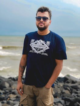 Portrait of a man in casual attire standing on a rocky beach with waves in the background.