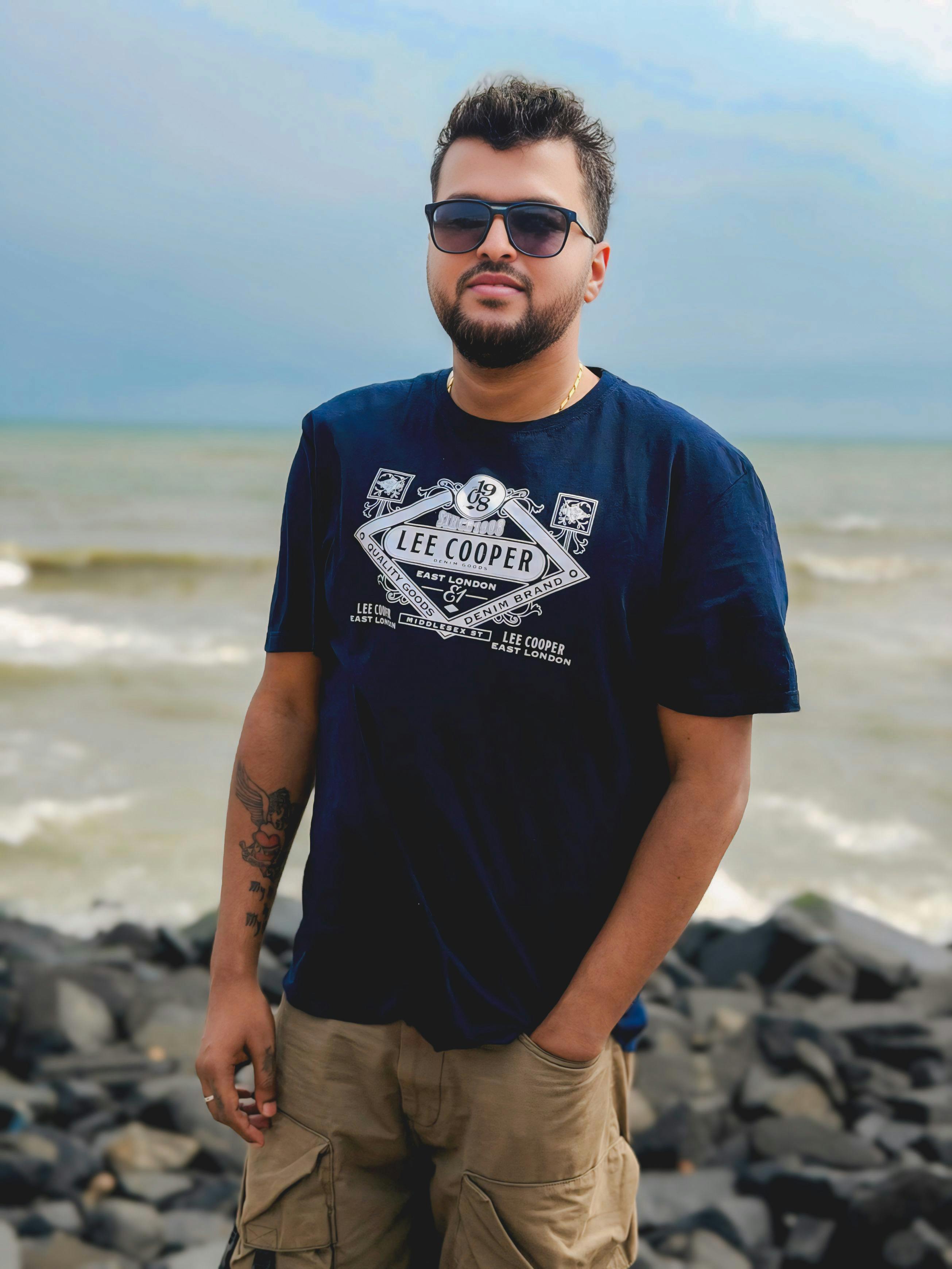 Gratis Retrato de un hombre vestido de manera informal, de pie en una playa rocosa con olas al fondo. Foto de stock