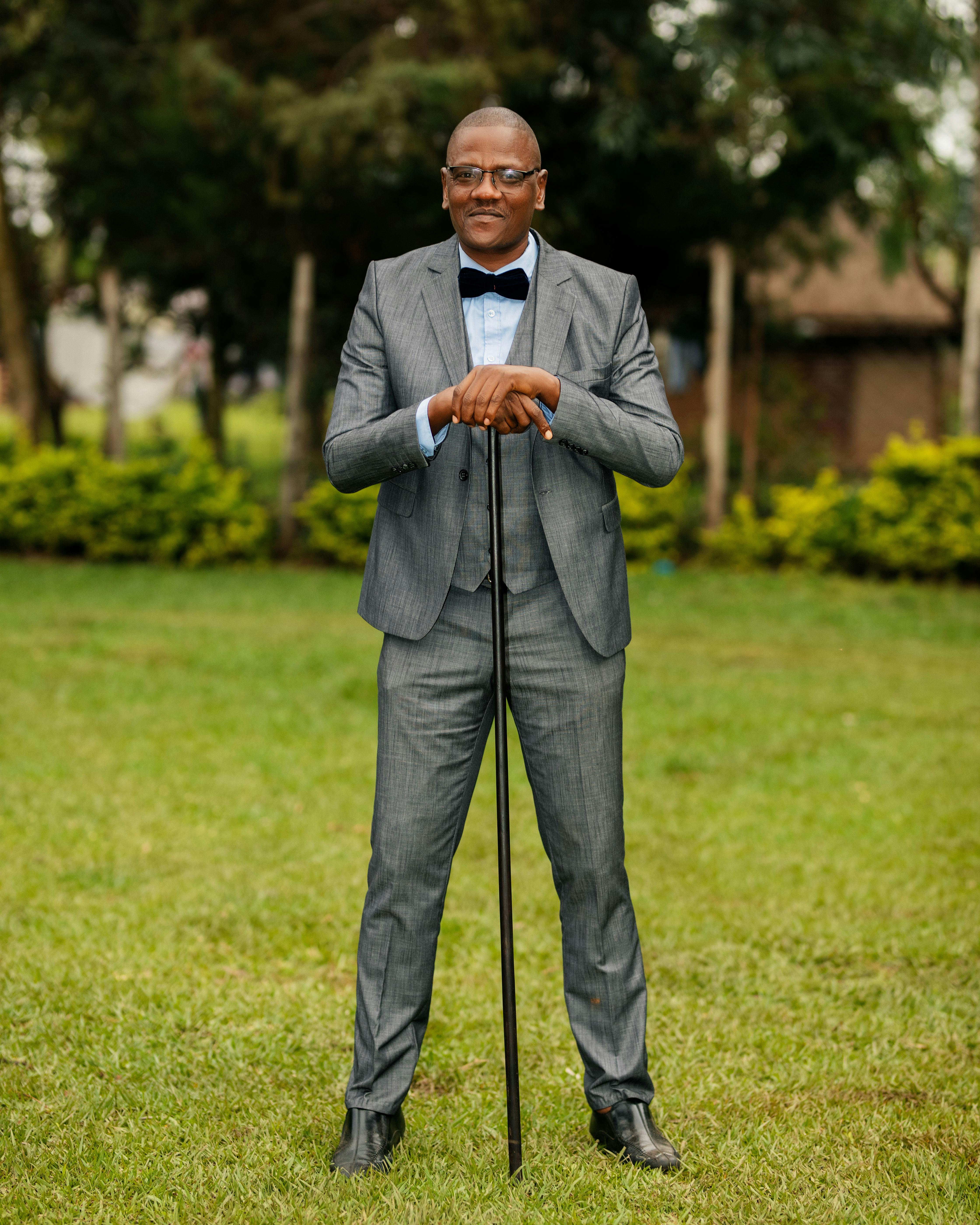 Stylish man in gray suit with cane standing in a lush garden setting.