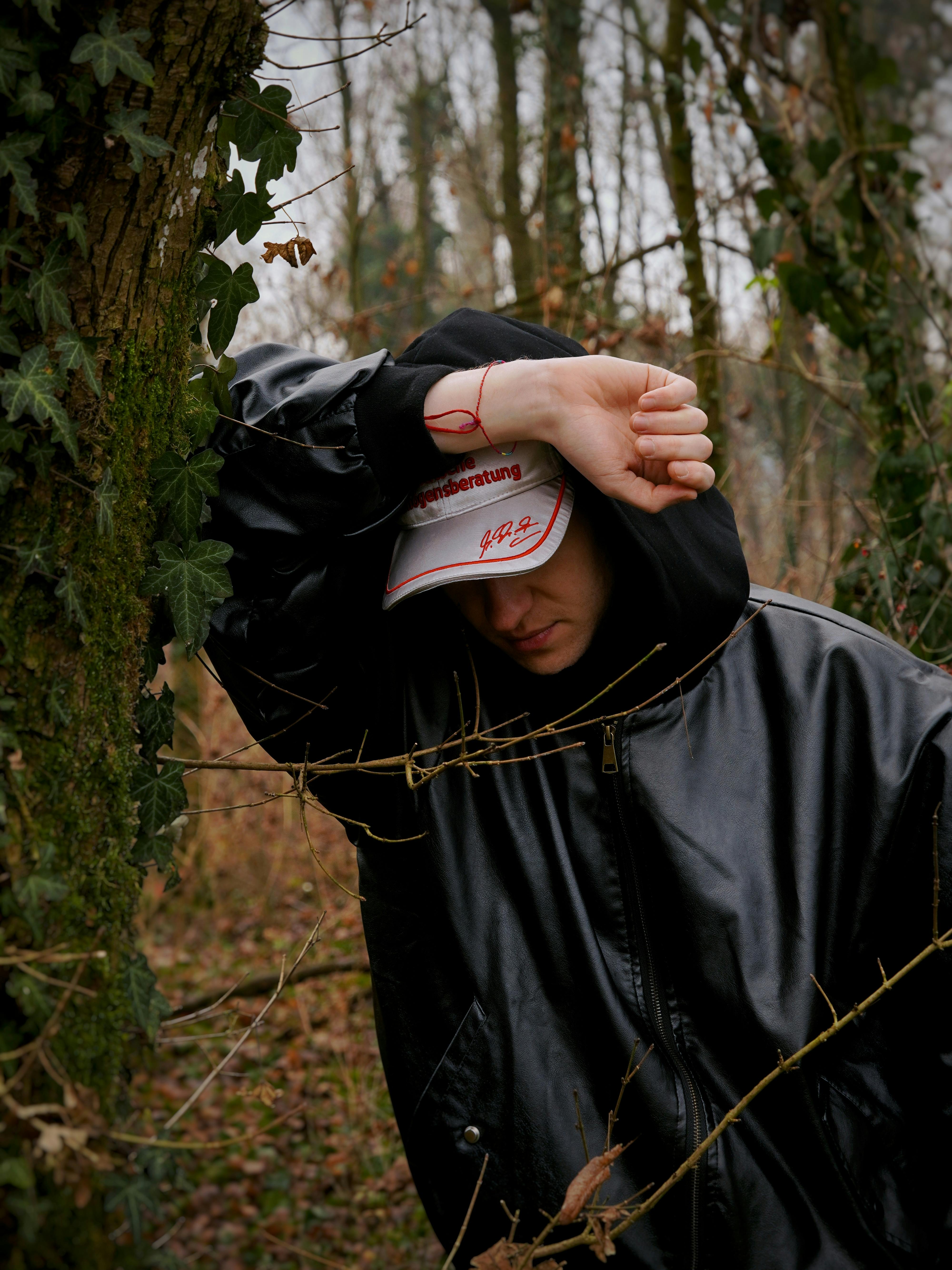 Person in a black leather jacket leans on tree in a forest setting.