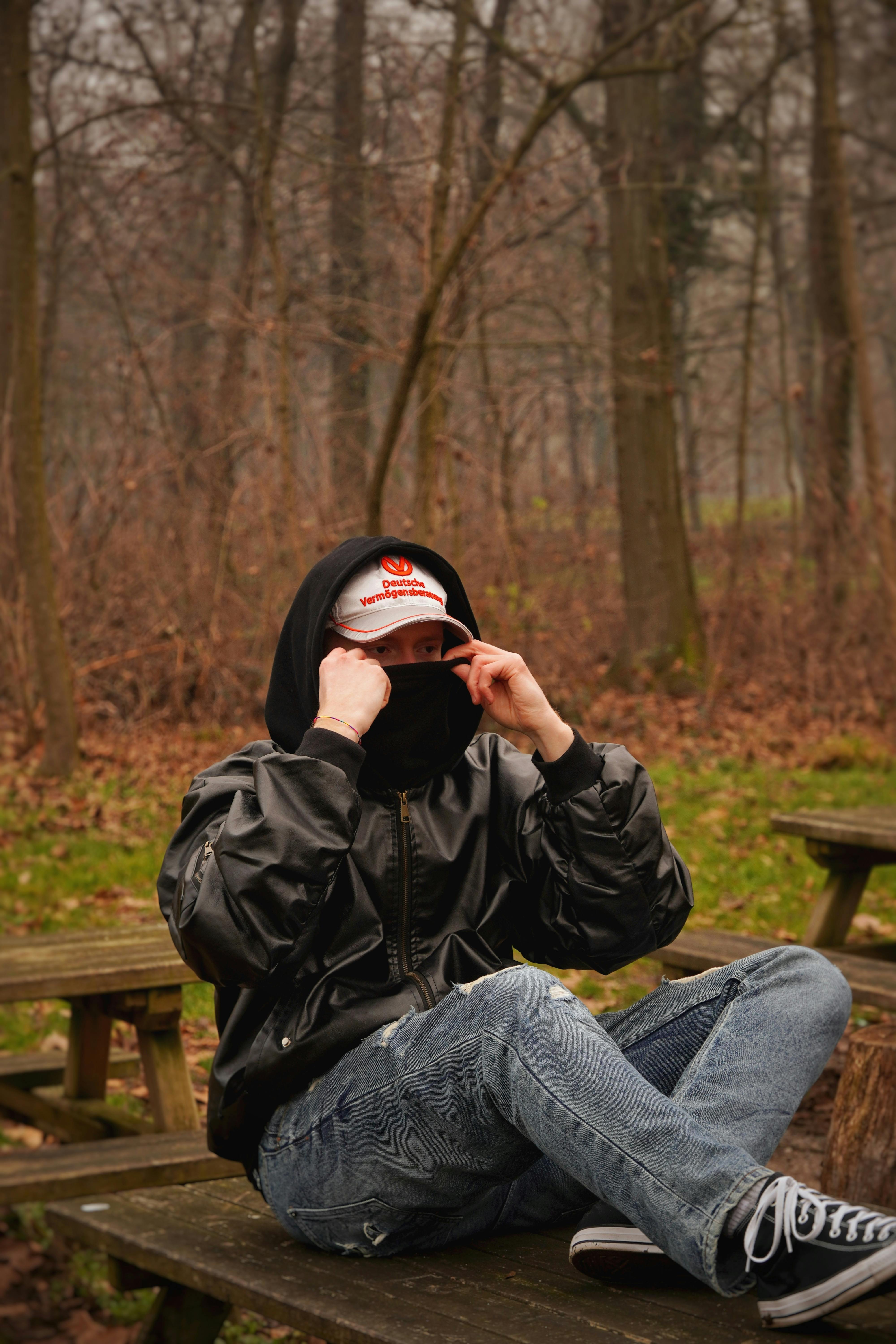 De franc Un adult jove amb una dessuadora amb caputxa i gorra seu en un banc en una zona boscosa, gaudint d'un moment de tranquil·litat. Foto d'estoc