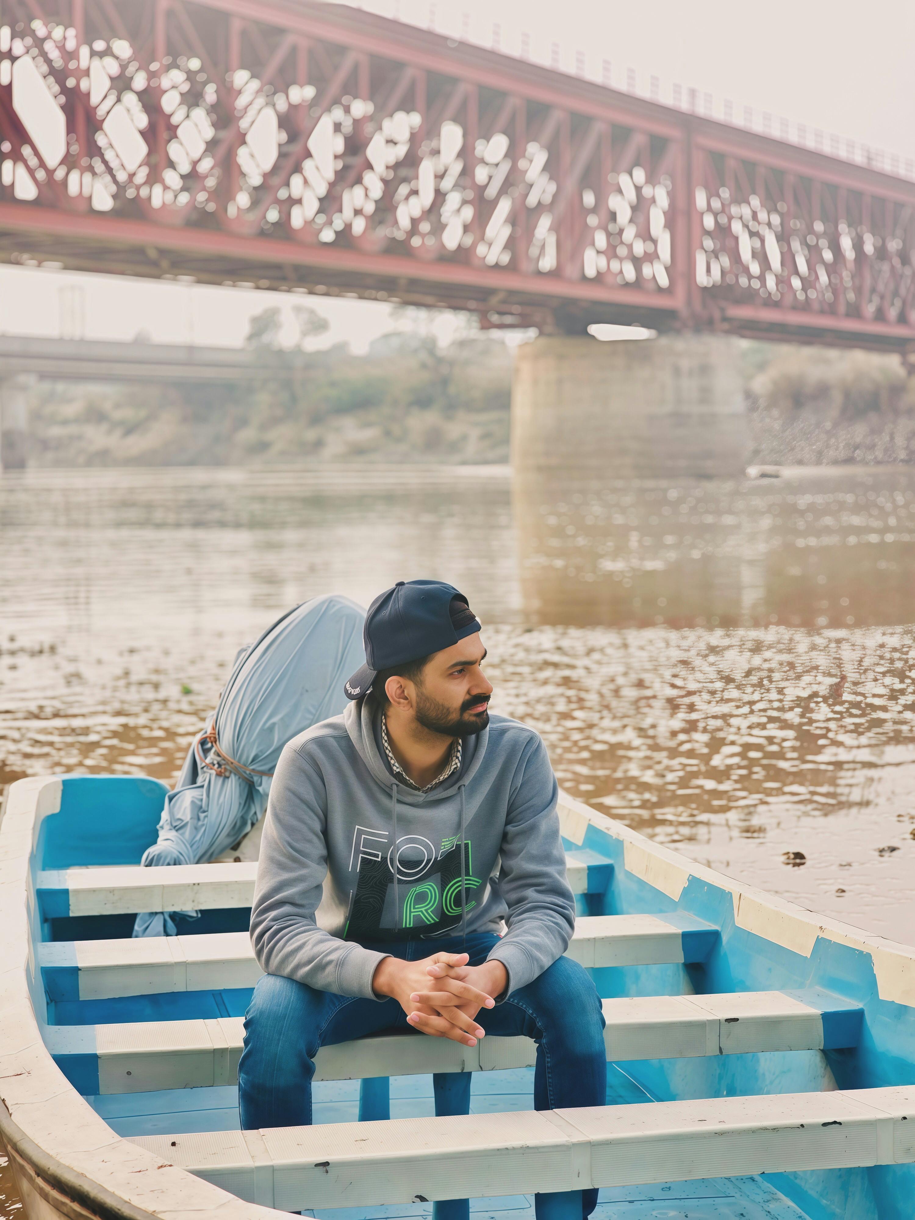 A young man sits in a boat by a river with a railway bridge in Faisalabad, Pakistan.