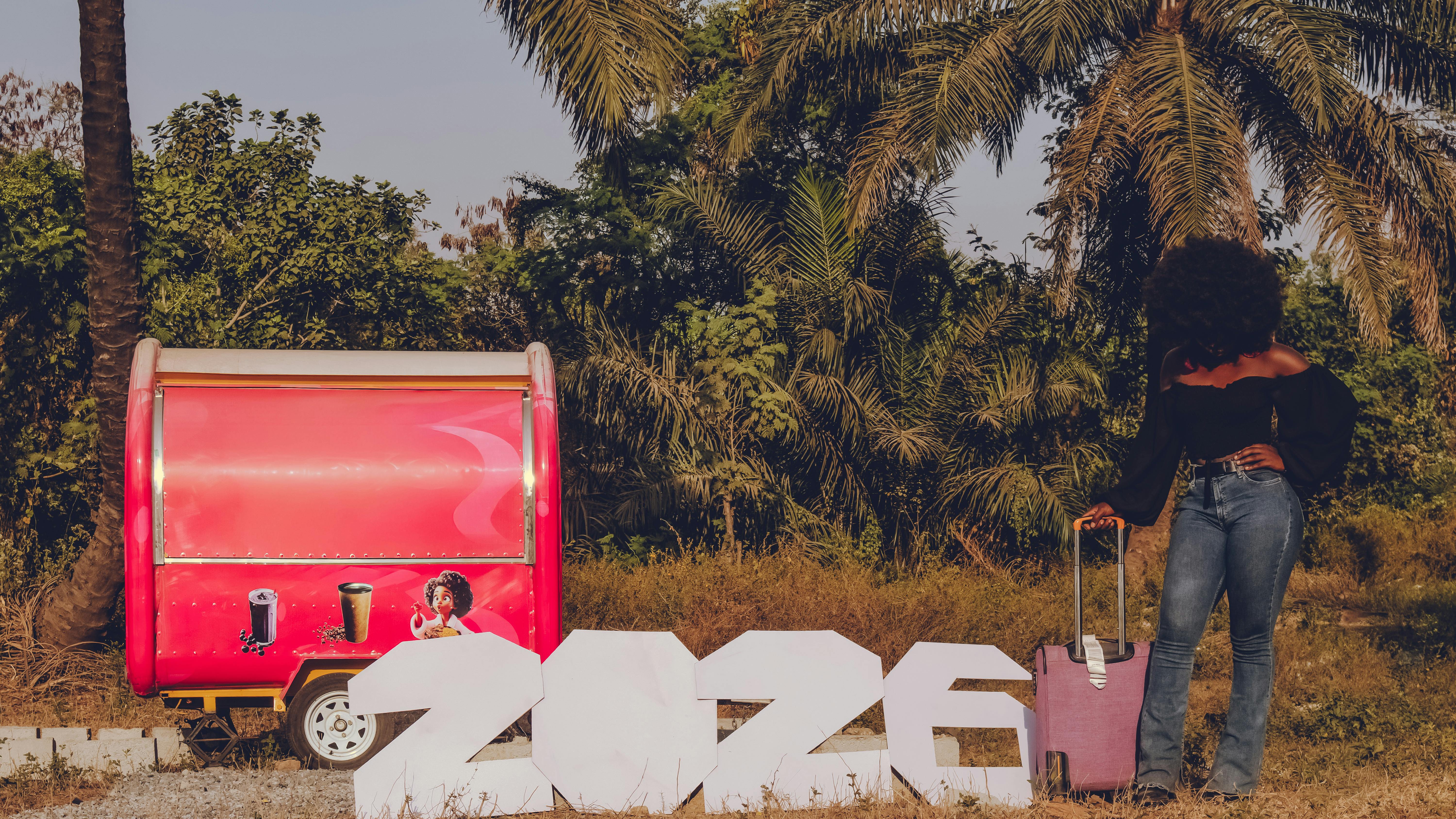 Gratis En kvinne med bagasje ved siden av et stort 2026-skilt i nærheten av en fargerik rød campingvogn utendørs. Arkivbilde