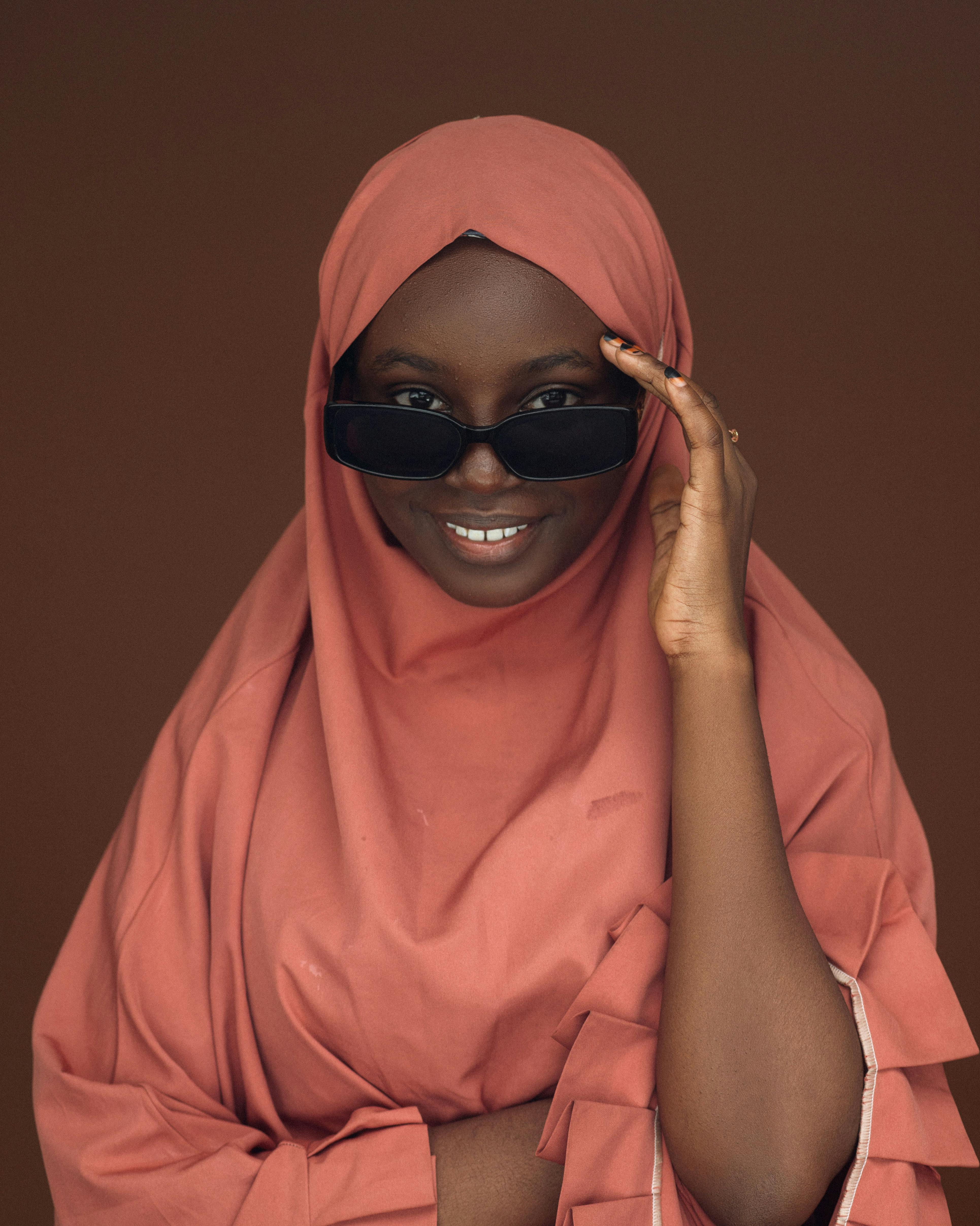 Gratis Retrato de una mujer con un hijab rosa, gafas de sol y sonriendo sobre un fondo marrón. Foto de stock