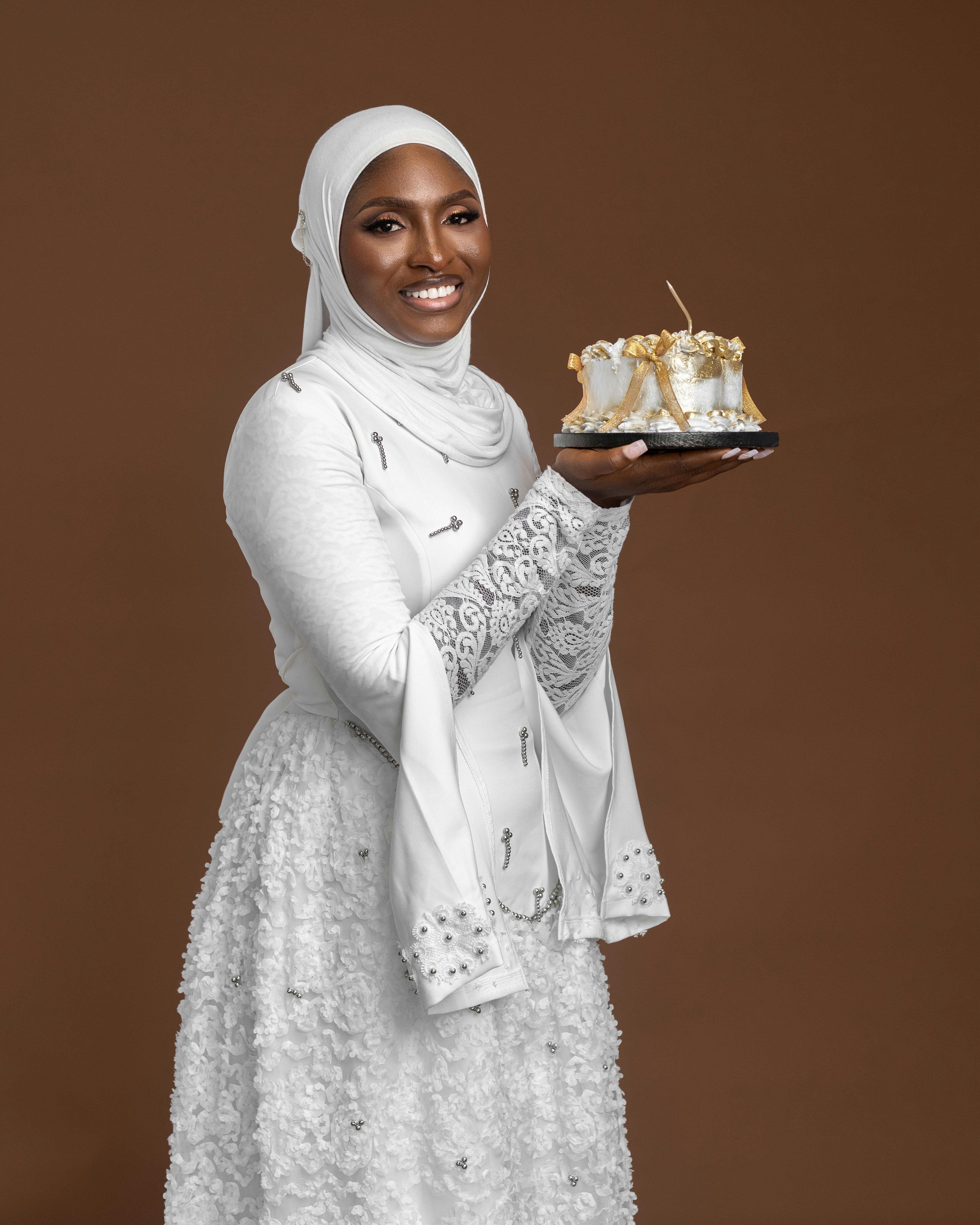 Gratis Mujer elegante con hijab blanco sosteniendo un pastel adornado sobre fondo marrón. Foto de stock