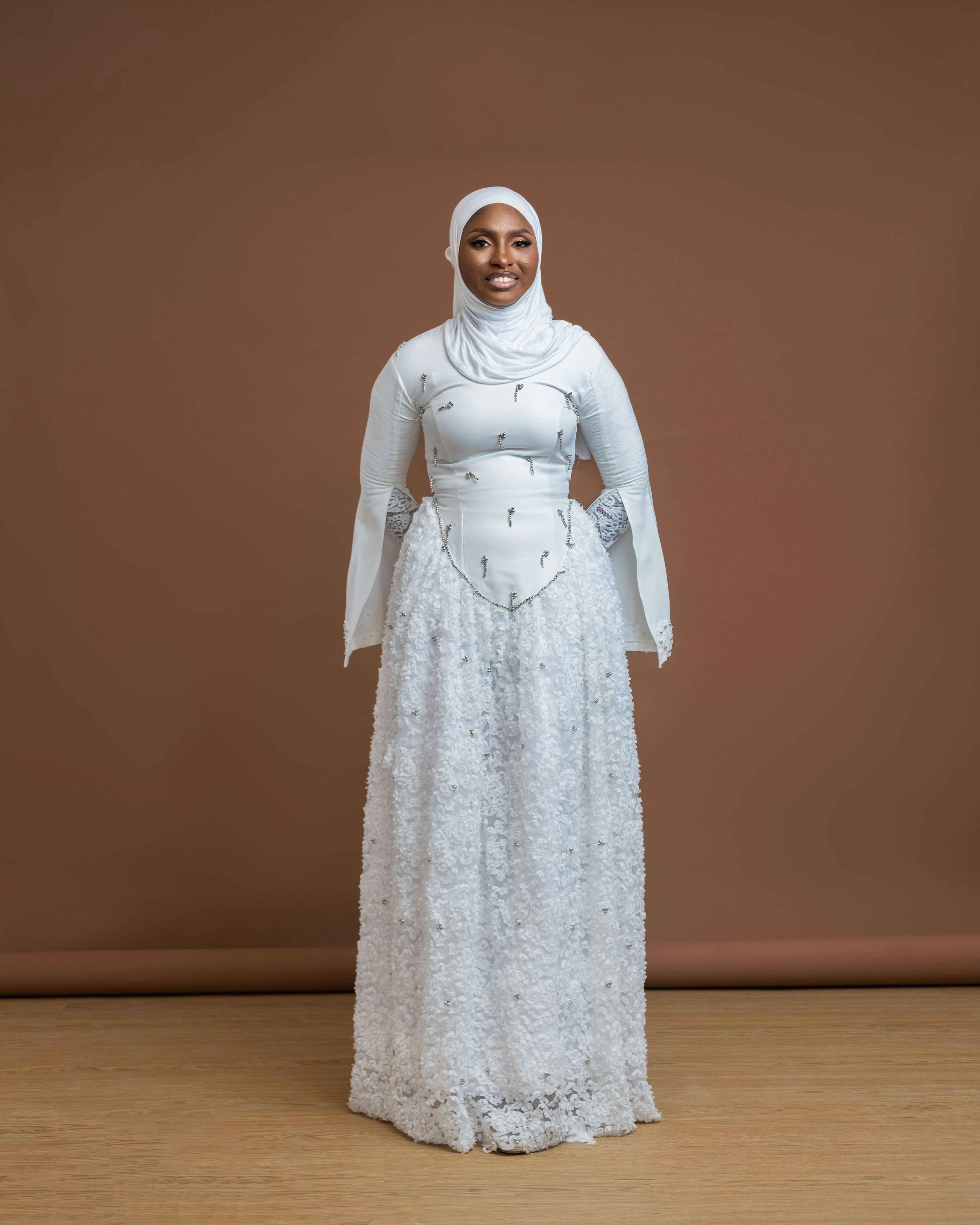 Gratis Retrato de una mujer sonriente con una bata blanca y un hijab, de pie sobre un piso de madera sobre un fondo marrón. Foto de stock