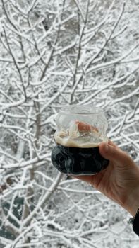 A hand holding a glass of dark beverage in front of a snowy winter landscape.