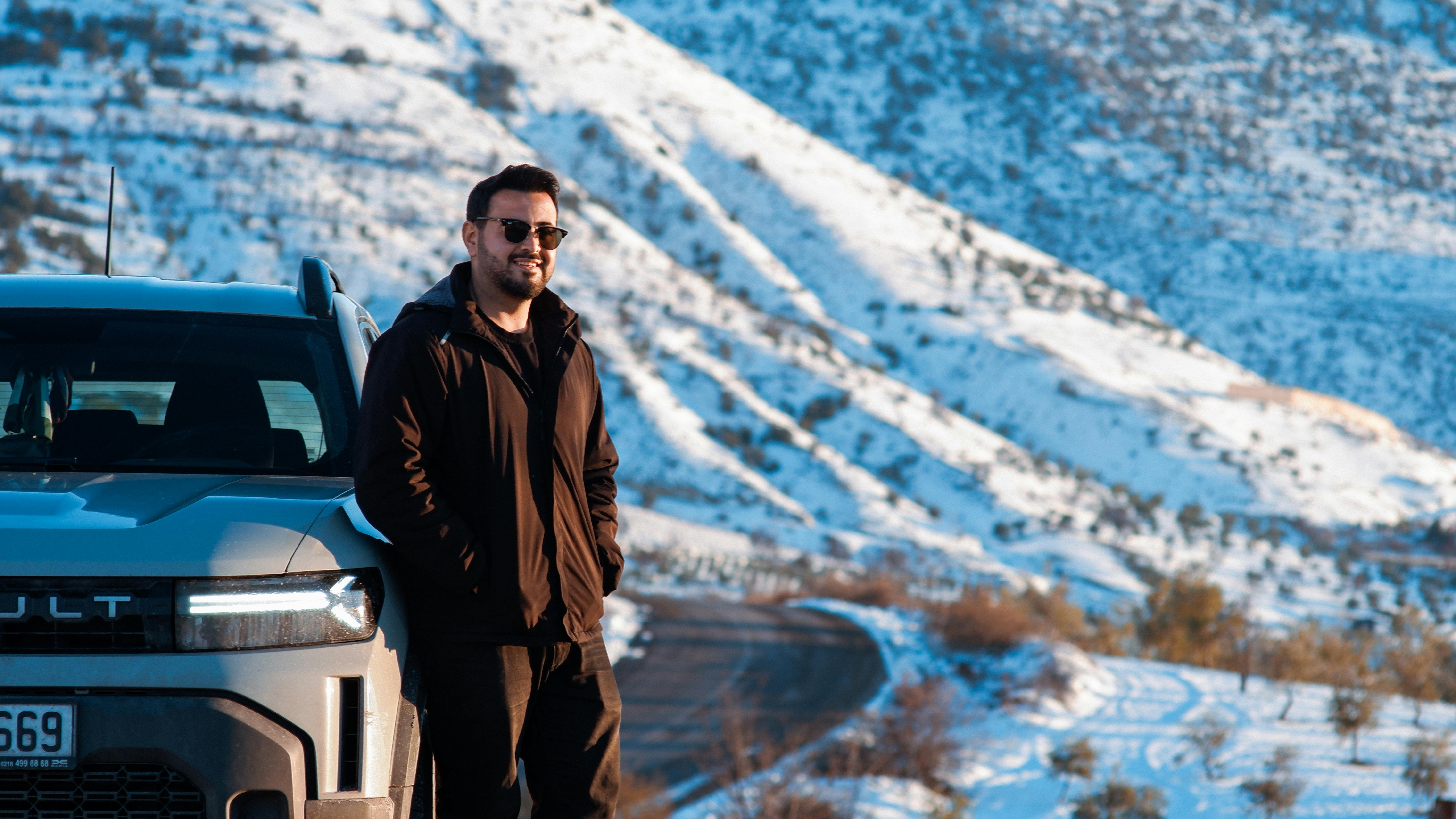Smiling man with sunglasses beside a car in scenic winter mountains.