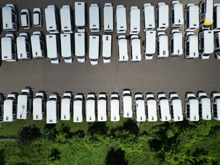 Photo by Bl∡ke Aerial view of a large parking lot filled with white vans on a sunny day.