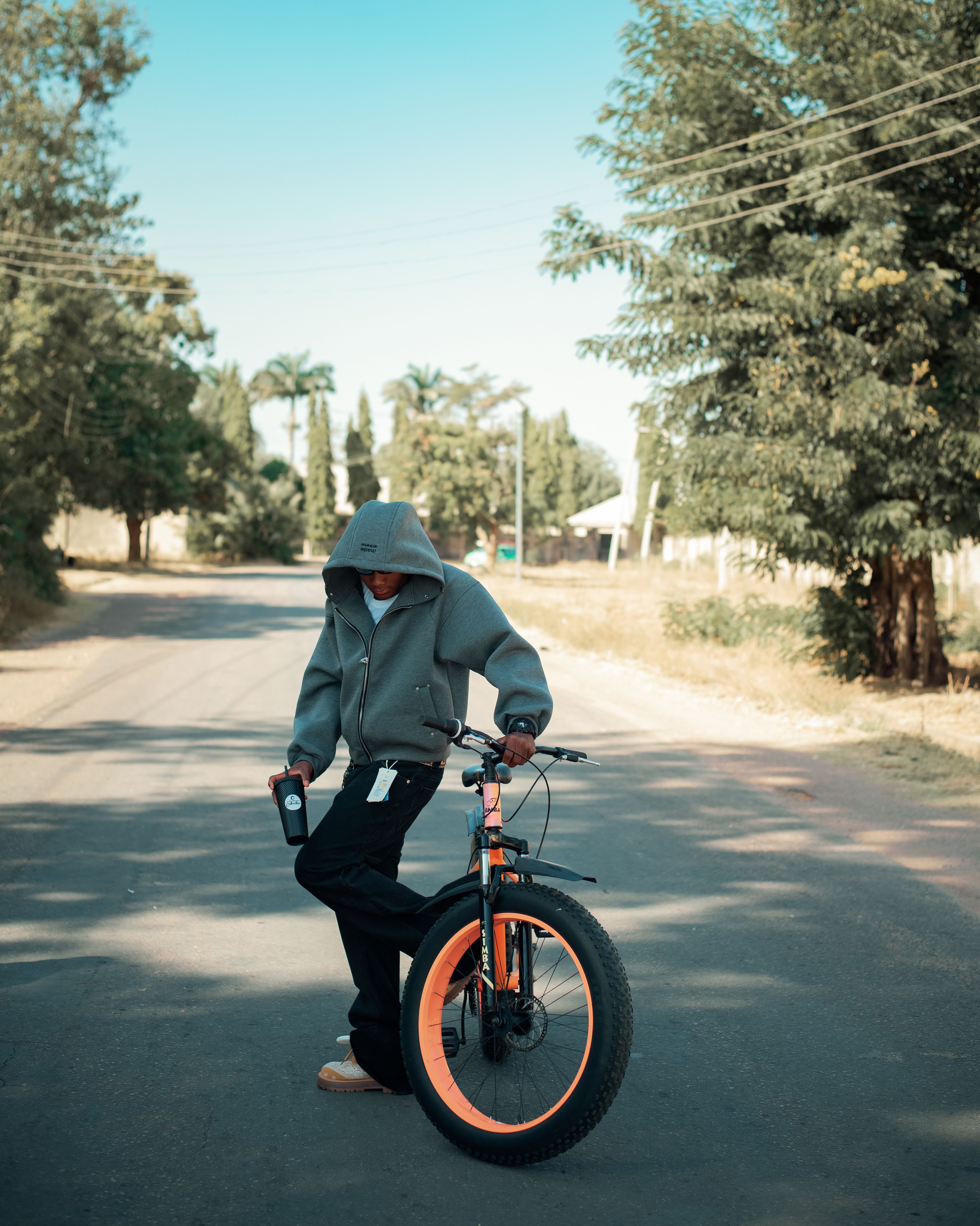 A teenager wearing a hoodie leans on a bicycle on a sunny rural road.