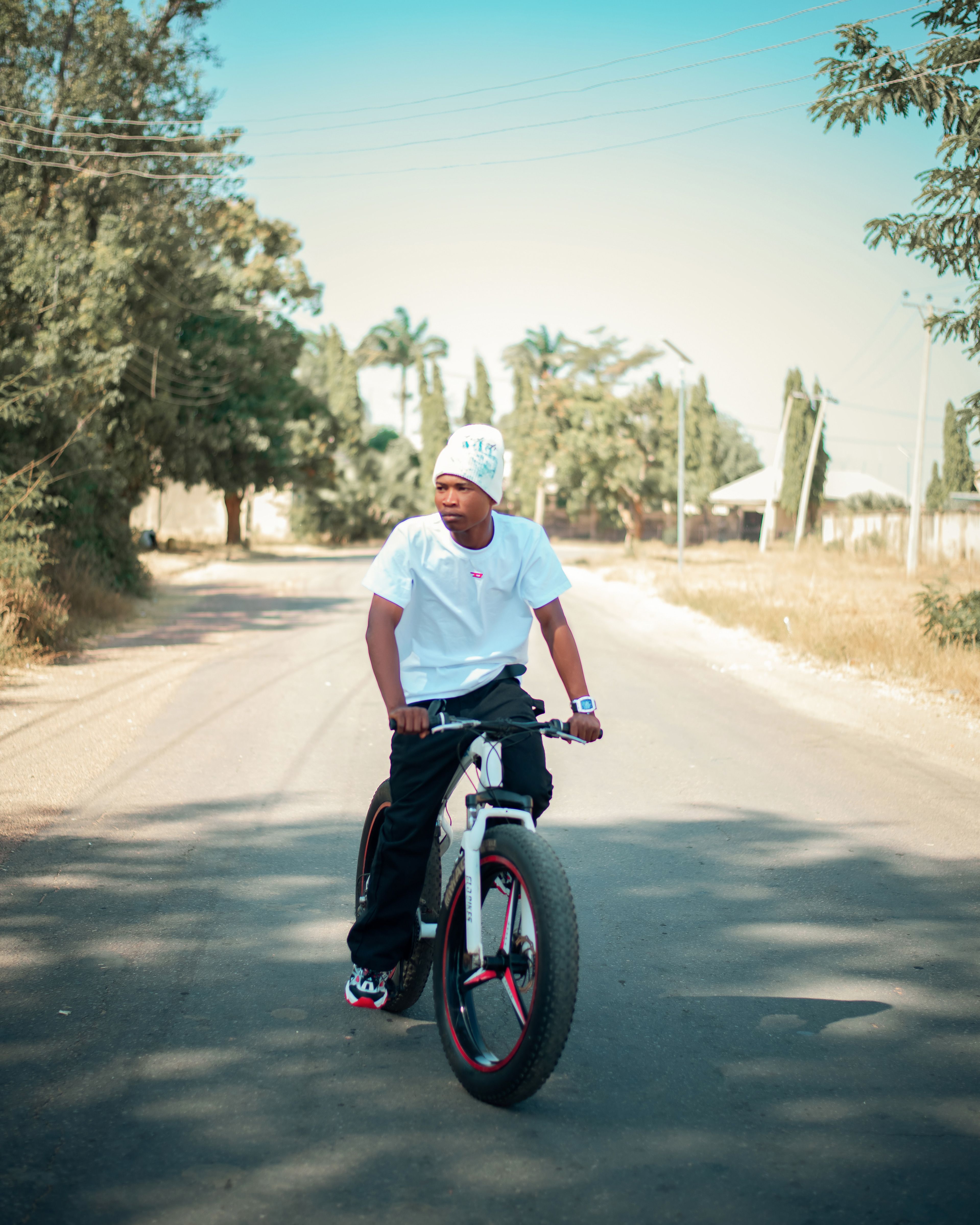Young man riding a bicycle on a sunny street, surrounded by trees and clear skies.