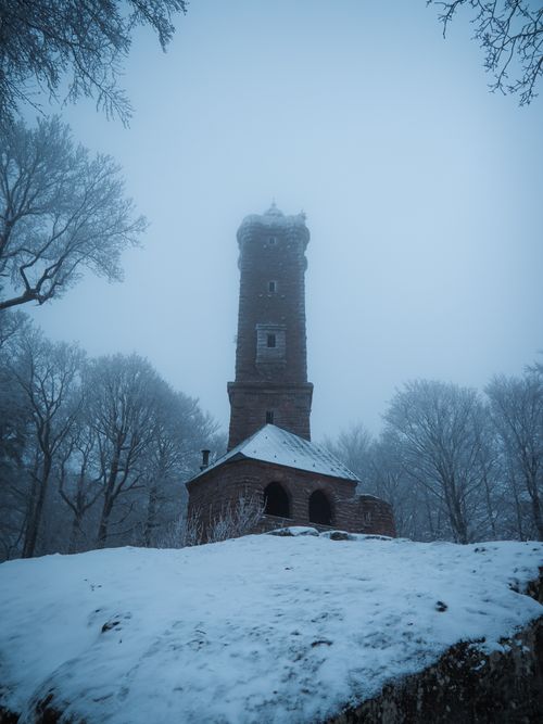 Free A tall, rustic tower stands amidst a snowy, mist-covered forest in winter. Stock Photo