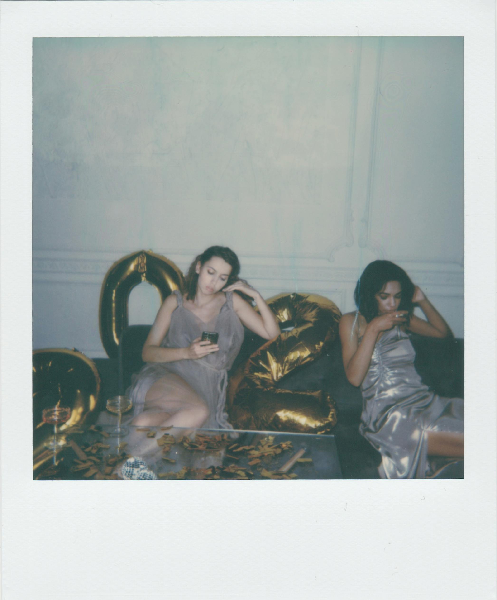 Two women in elegant dresses relaxing indoors with a vintage setting, surrounded by gold decor and balloons.