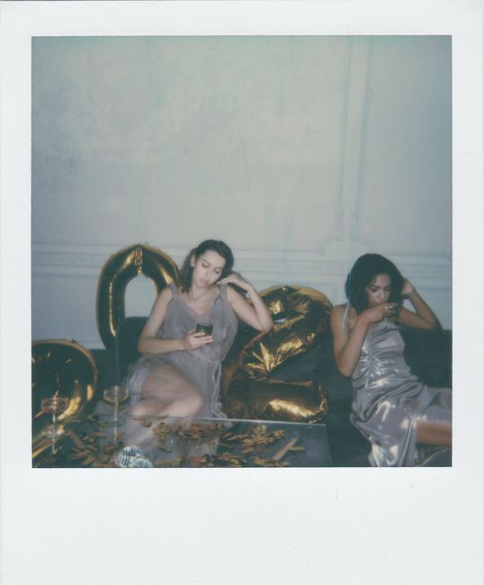 Two women in elegant dresses relaxing indoors with a vintage setting, surrounded by gold decor and balloons.