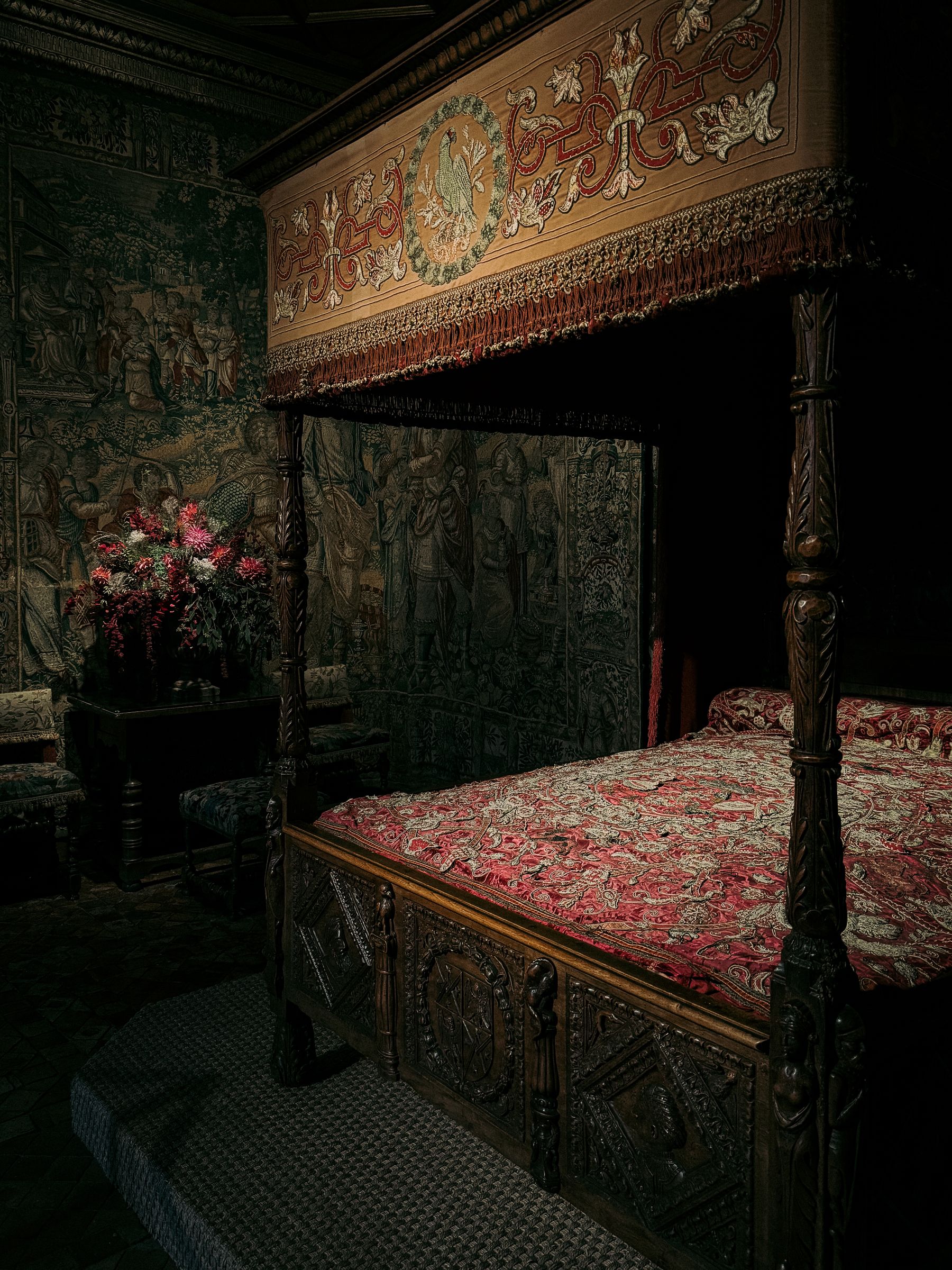 Dark carved canopy bed with red bedding, patterned tapestries and exposed timber in a historic bedroom