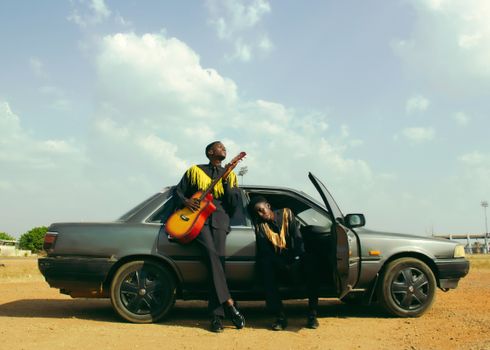 Two stylish musicians with a guitar sitting by a classic car under a bright sky.