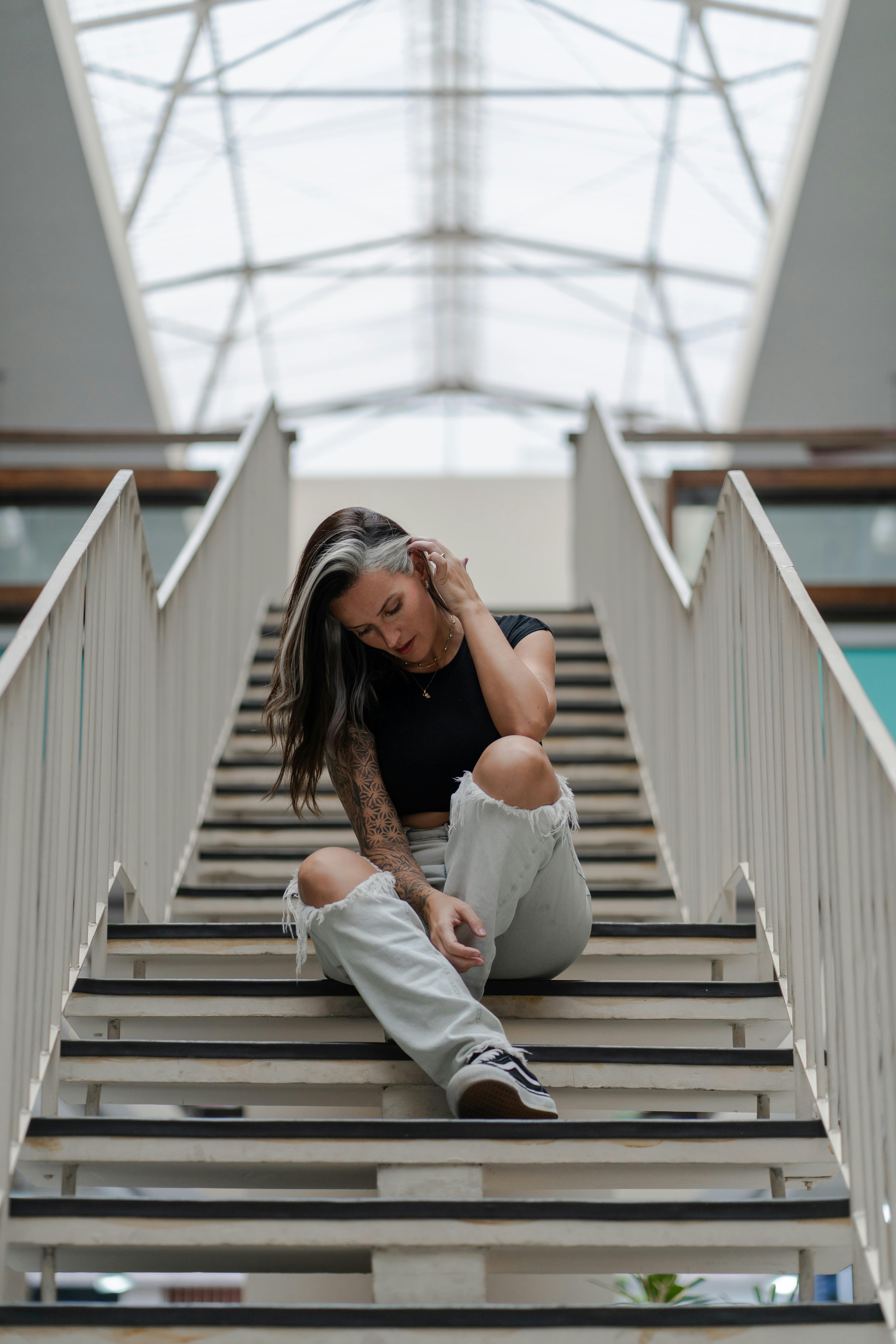De franc Una dona jove amb roba informal seu pensativa a l'escala interior amb vistes arquitectòniques al sostre. Foto d'estoc