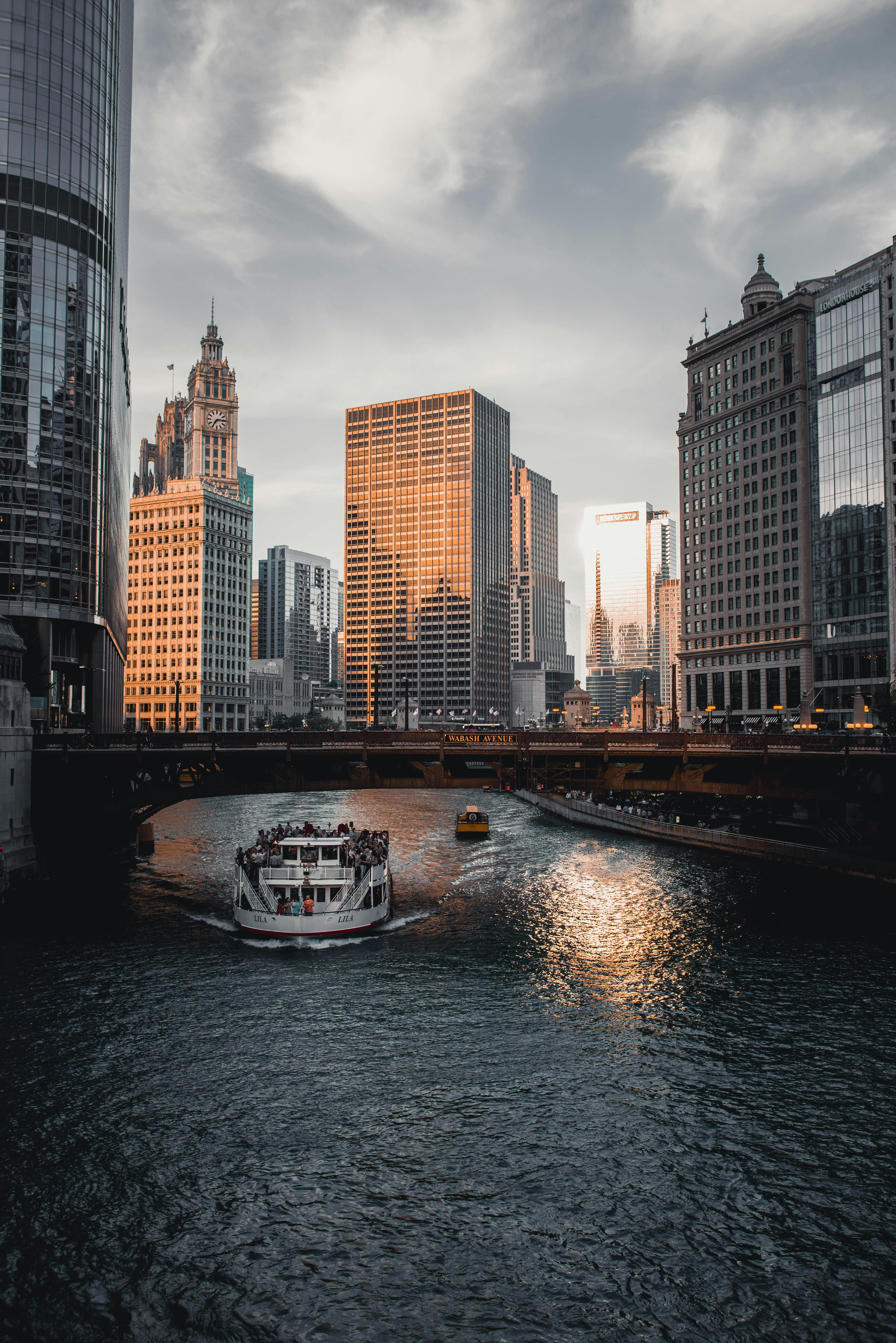 日落时分，一艘河船在桥下航行，映衬着城市的天际线，展现了城市生活与建筑的景象