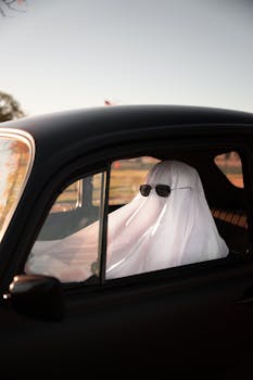 A ghost covered in a sheet wearing sunglasses in a vintage car, outdoors.