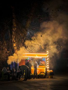 An atmospheric nighttime scene of a street food stall glowing with warm lights and surrounded by people.