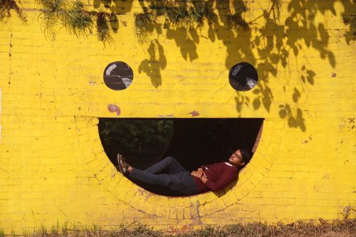 Free A man rests inside a smiley face cutout on a bright yellow wall under sunlit shadows. Stock Photo
