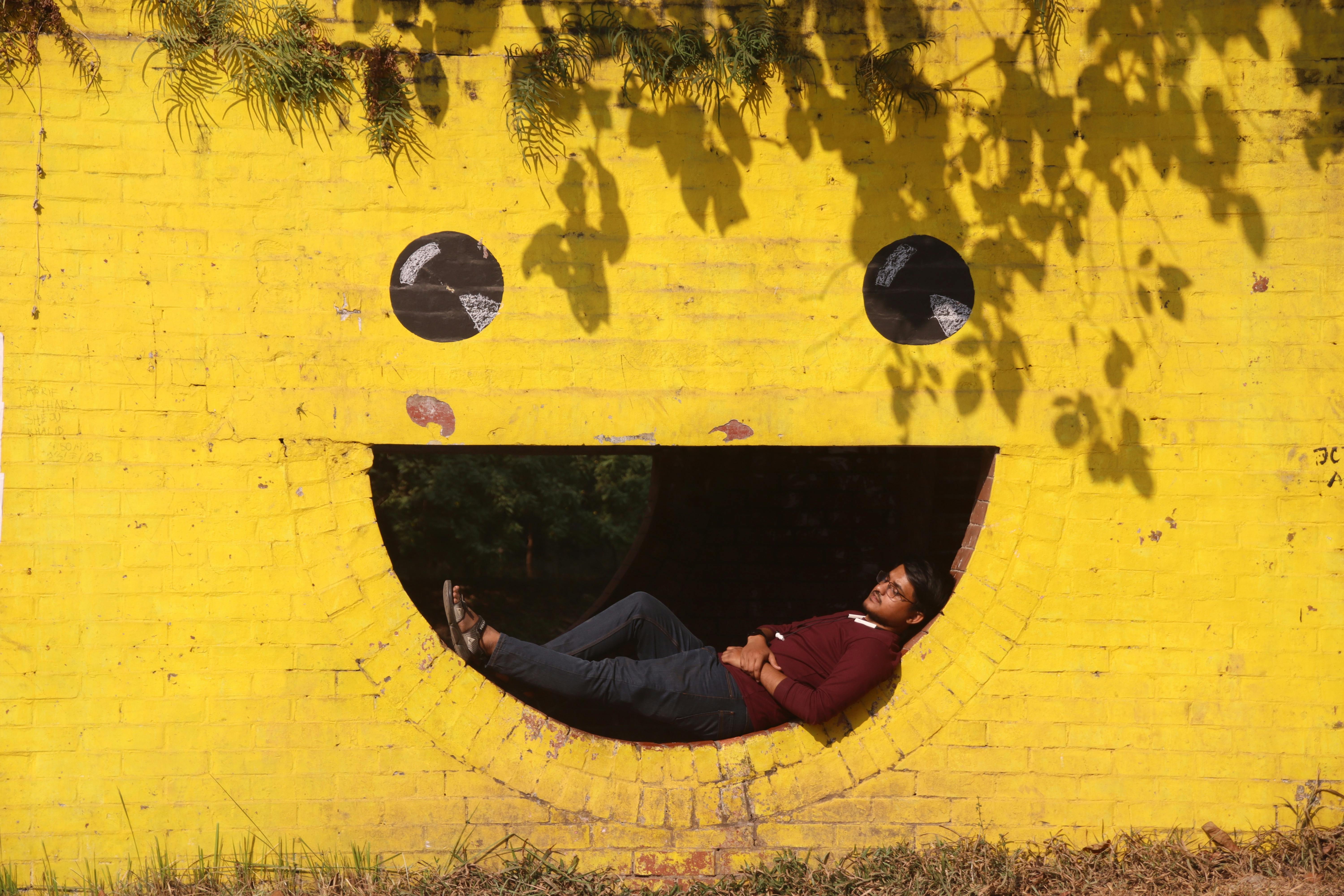 Gratis En mann hviler inni et smilende ansikt på en knallgul vegg under solbelyste skygger. Arkivbilde