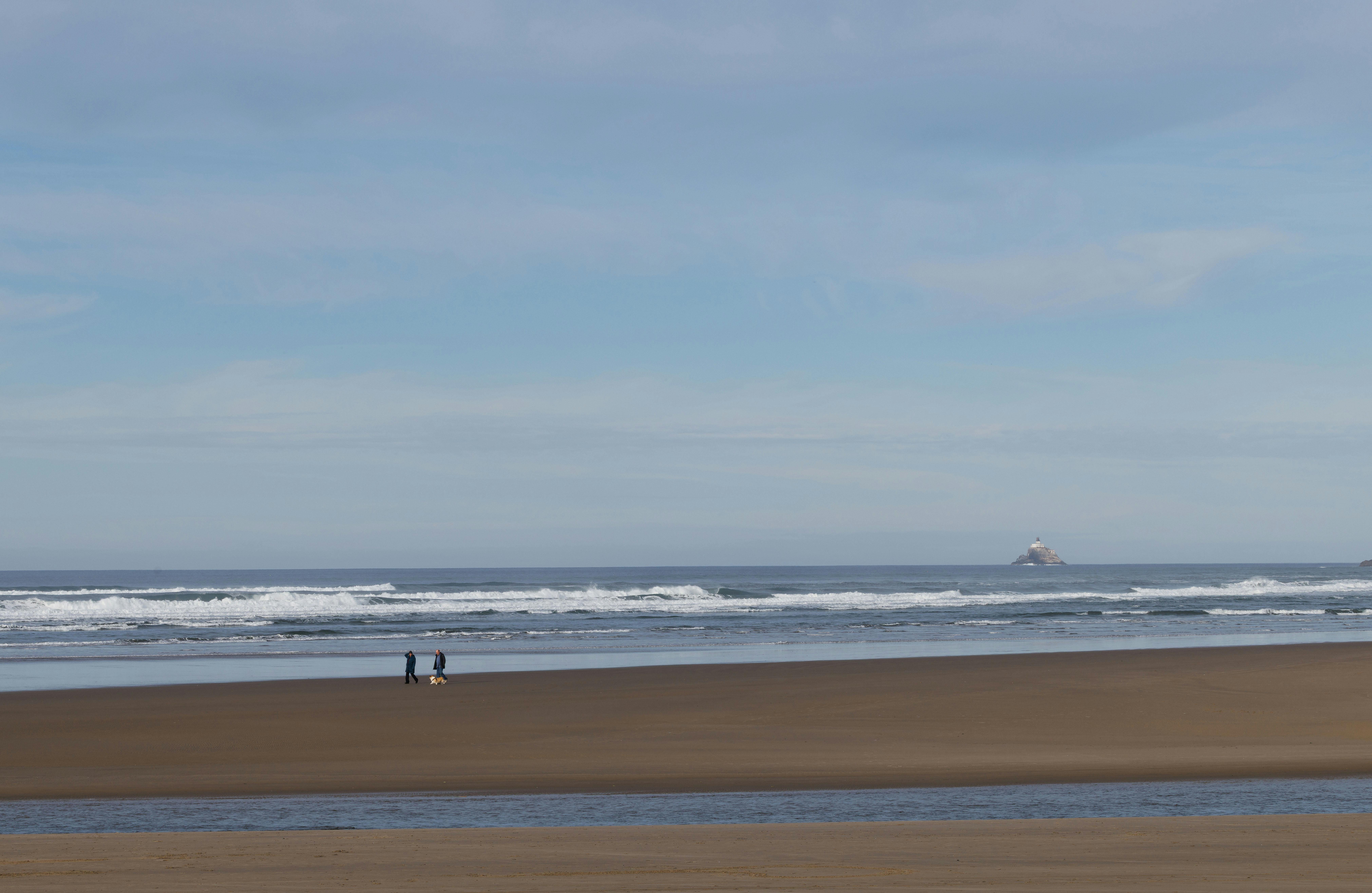 Tranquil beach scene with two people walking by the ocean, distant lighthouse visible.
