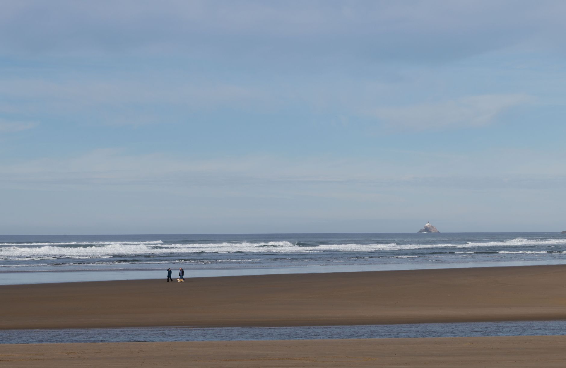 Tranquil beach scene with two people walking by the ocean, distant lighthouse visible.