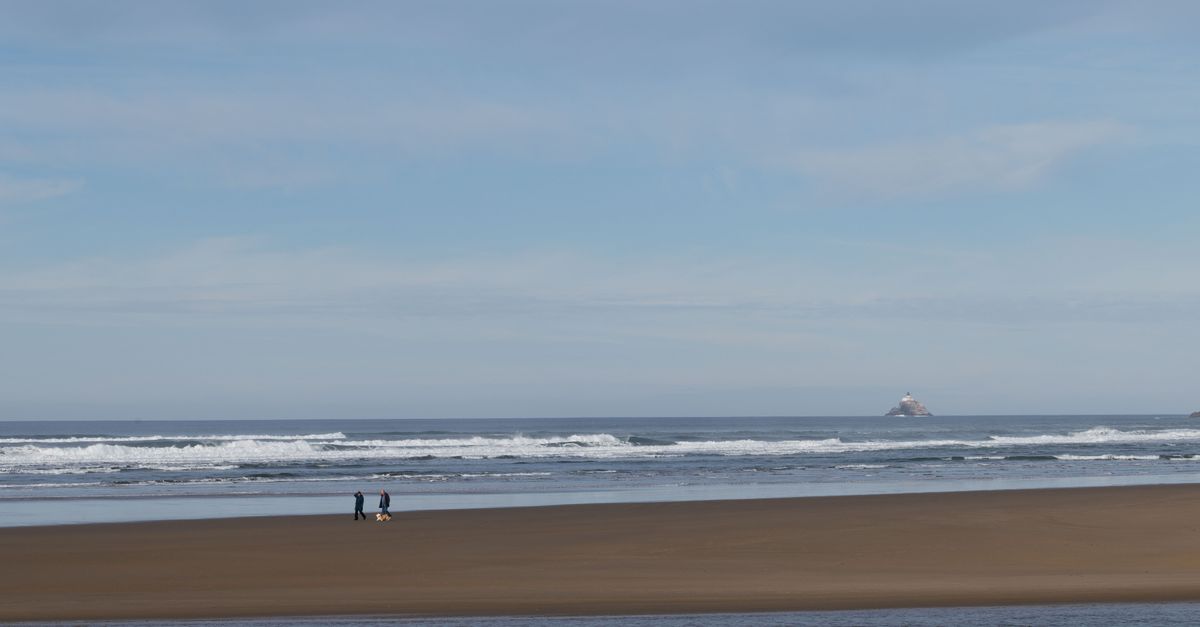 Tranquil beach scene with two people walking by the ocean, distant lighthouse visible.