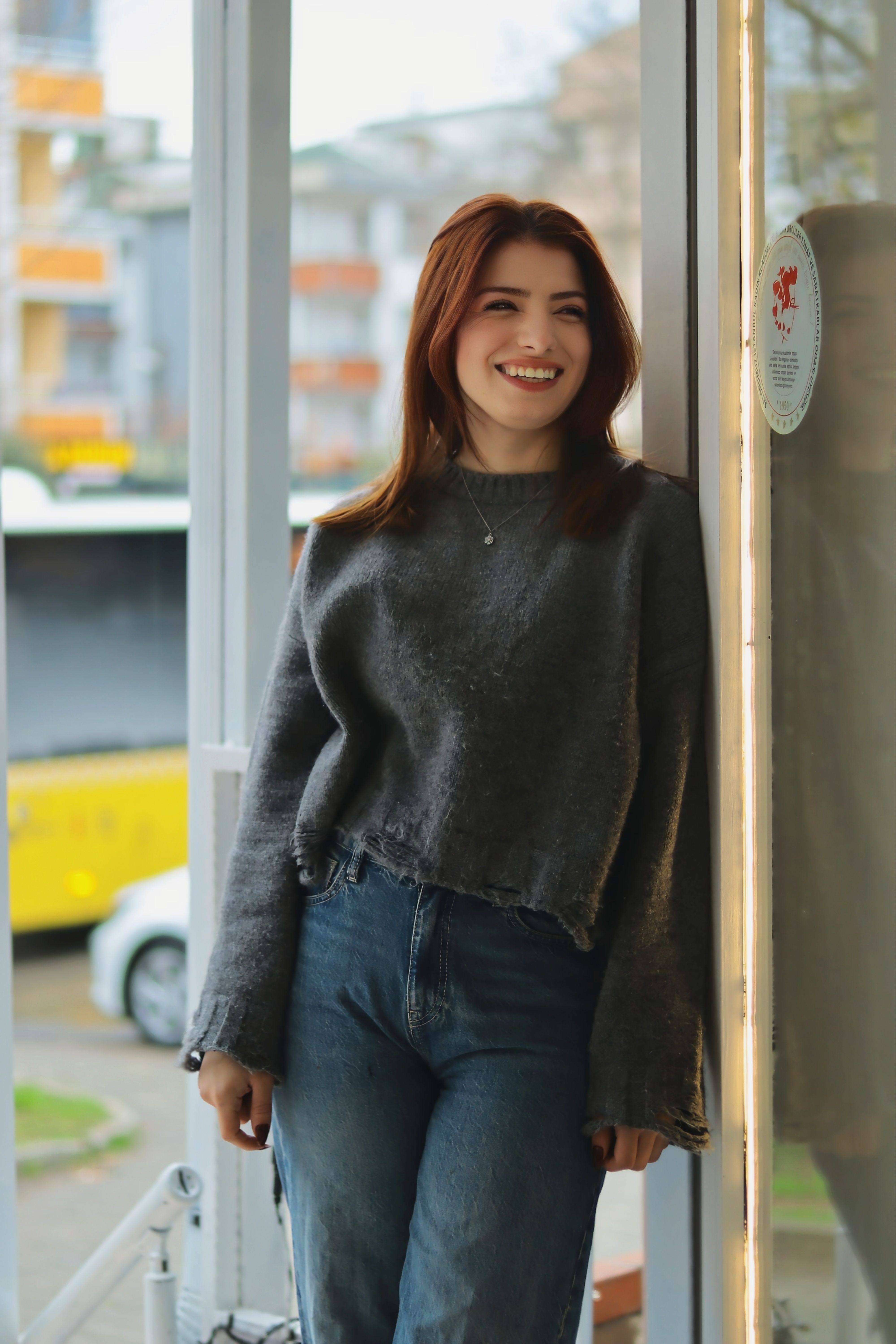 Smiling young woman leaning against a window in an urban setting, casual fashion.
