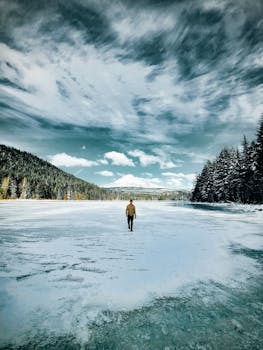 A lone person walks on a vast frozen lake surrounded by snow-covered trees.