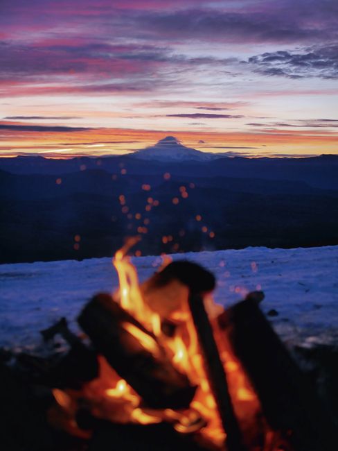 A warm campfire overlooking a majestic mountain silhouette against a colorful sunset sky.
