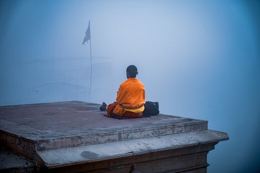 A monk in orange robes meditates peacefully in the morning mist by the Ganges River in Varanasi.