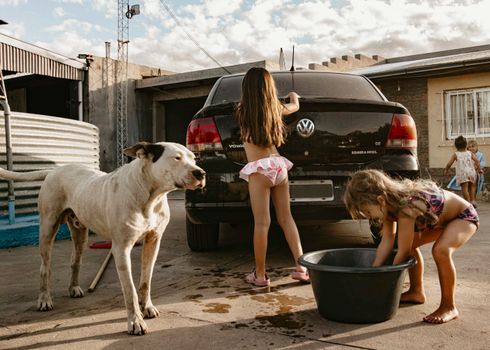 Two children wash a car in a sunny backyard with a dog nearby, creating a warm summer scene.