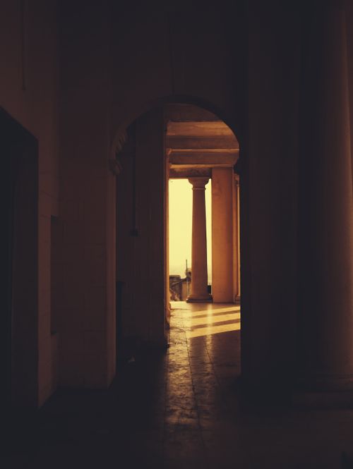 Free A warm, sunlit corridor with classical pillars casts shadows in an ancient building. Stock Photo