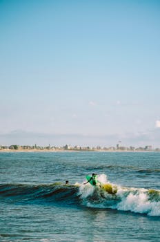 Surfer catching a wave near a coastline with urban skyline, blue sky overhead.