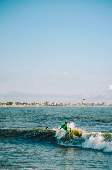 Surfer catching a wave near a coastline with urban skyline, blue sky overhead.