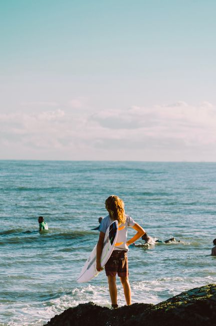 Young surfer holding a surfboard, looking at the ocean waves on a bright summer day.
