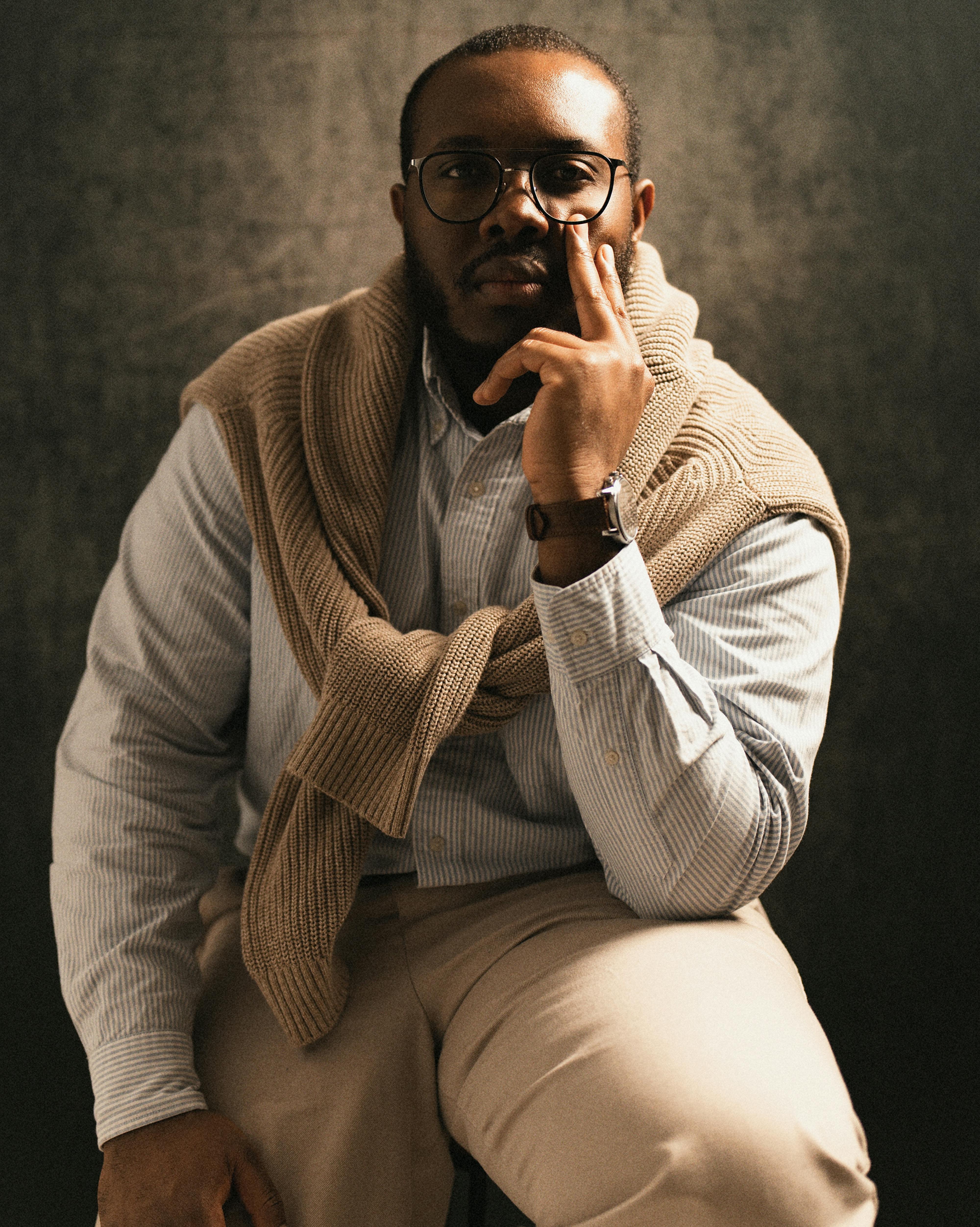 A sophisticated adult man posing confidently wearing glasses and a sweater indoors.